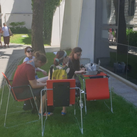 Four people are sitting at a computer in the garden of ms1. The computer stands on a small desk. A museum employee shows guests the portal zasoby.msl.org.pl