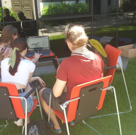 Four people are sitting at a computer in the garden of ms1. The computer stands on a small desk. A museum employee shows guests the portal zasoby.msl.org.pl
