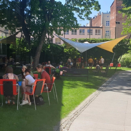 Four people are sitting at a computer in the garden of ms1. The computer stands on a small desk. A museum employee shows guests the portal zasoby.msl.org.pl. In the background is the museum garden with other guests