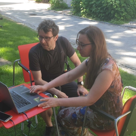 Two people are sitting at a computer in the garden ms1. The computer stands on a small desk. A museum employee shows guests the portal zasoby.msl.org.pl