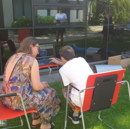 Two people are sitting at a computer in the garden ms1. The computer stands on a small desk. A museum employee shows guests the portal zasoby.msl.org.pl
