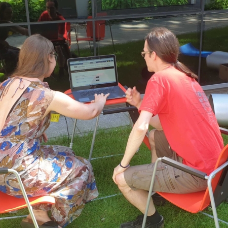 Two people are sitting at a computer in the garden ms1. The computer stands on a small desk. A museum employee shows guests the portal zasoby.msl.org.pl