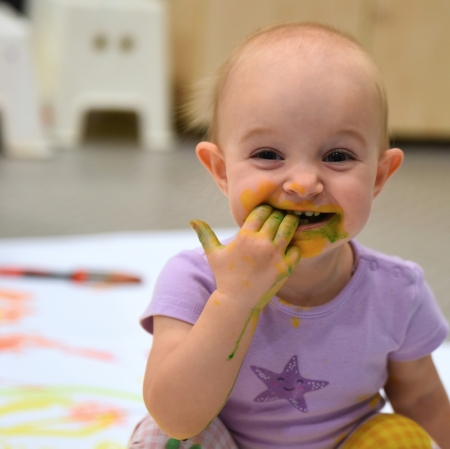 a girl holding her hand in her mouth, the child's hand and face are covered in paint