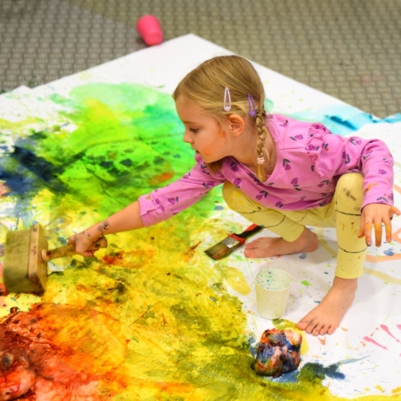 little girl sitting on cardboard painted with splashes of colorful paints and painting with a large paint brush