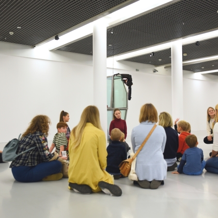 a group of adults and children are sitting on the ground in the exhibition space. In the background you can see glass sculptures by the artist Agata Ingarden