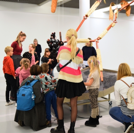 a group of adults and children in the exhibition space listening to an educator showing around the exhibition