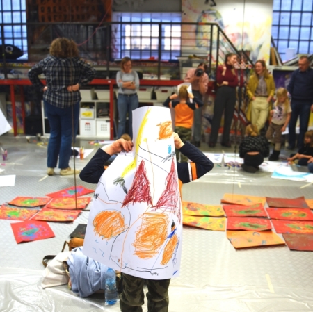 a child covering himself with a cardboard with his work, standing in the space of a workshop room