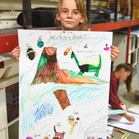 a girl holding her work in front of her, which includes a dinosaur, a volcano, a cat and many other characters
