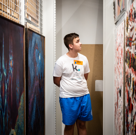 A young man dressed in a white T-shirt and blue shorts stands in the middle of the partitions on which paintings hang
