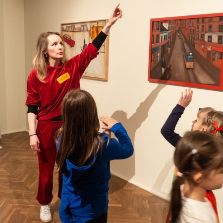 Exhibition space. Children face the person who points to the picture on the wall.