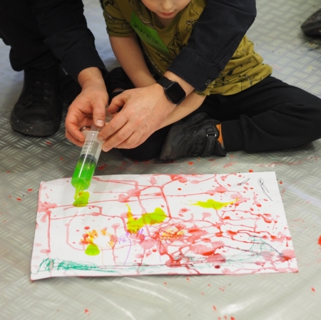 The child sits on the floor. In front of him is a piece of paper stained with paint. The man holds a syringe with paint in his hands.