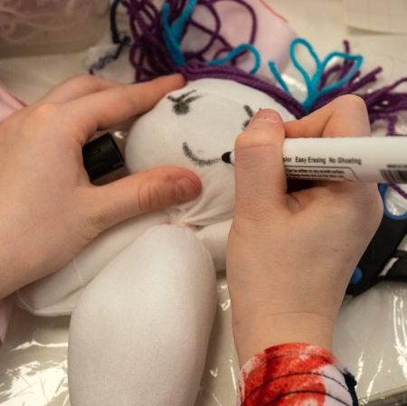 A close-up of a child's hands drawing a smile on a handmade doll with a marker.