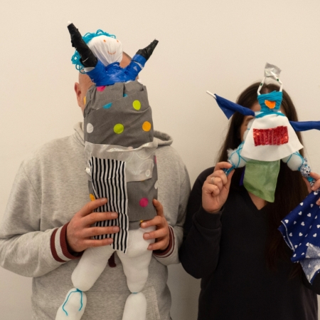 Two people stand next to each other. In their hands, at head level, they hold handmade dolls.