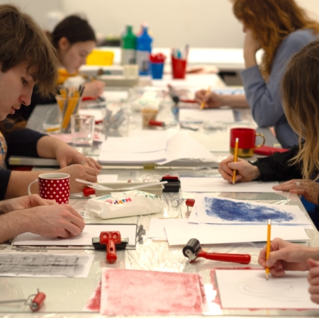 a group of people at a long table with a lot of sheets of paper and painting tools on it