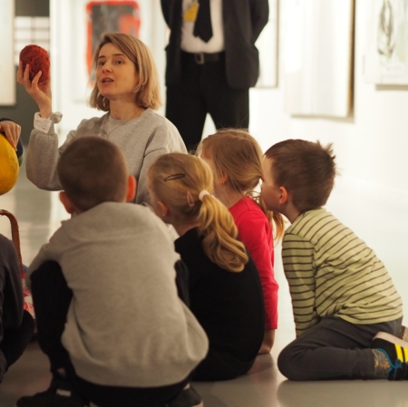 Children are sitting on the floor, visible from behind. In front of them is a woman. In the hand with which he raises the yarn
