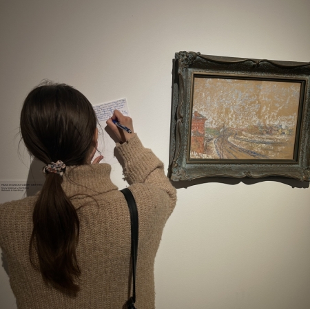 the girl, seen from the back, puts a piece of paper against the wall and writes on the paper. to her right is a painting