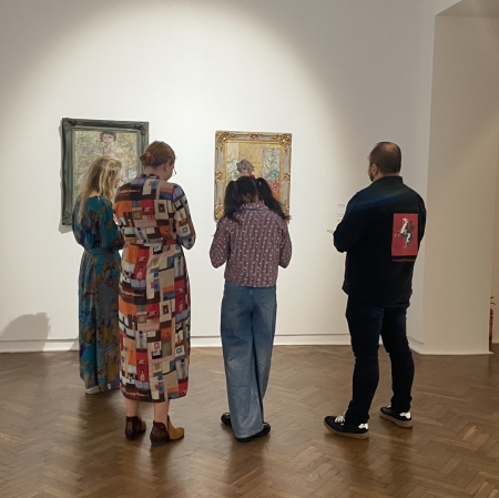 a group of people of different ages in the exhibition space listening to the educator