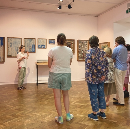 kilka osób w przestrzeni wystawy, patrzą na osobę znajdującą się przed nimi 