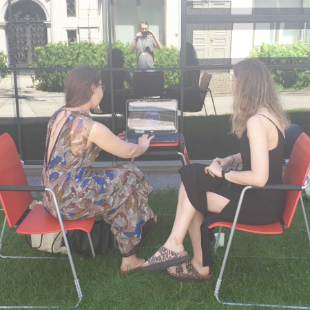Two people are sitting at a computer in the garden ms1. The computer stands on a small desk. A museum employee shows guests the portal zasoby.msl.org.pl