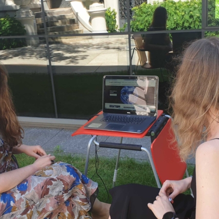 Two people are sitting at a computer in the garden ms1. The computer stands on a small desk. A museum employee shows guests the portal zasoby.msl.org.pl