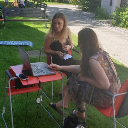 Two people are sitting at a computer in the garden ms1. The computer stands on a small desk. A museum employee shows guests the portal zasoby.msl.org.pl