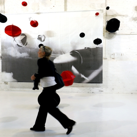 Osoba w przestrzeni wystawy, w tle czarno-białe duże zdjęcie wiszące na ścianie oraz kuliste, czerwone i czarne obiekty