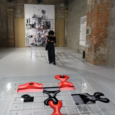 Osoba w przestrzeni wystawy, w tle czarno-białe zdjęcie wiszące na ścianie oraz obiekty w kolorze czerwonym, czarnym i białym