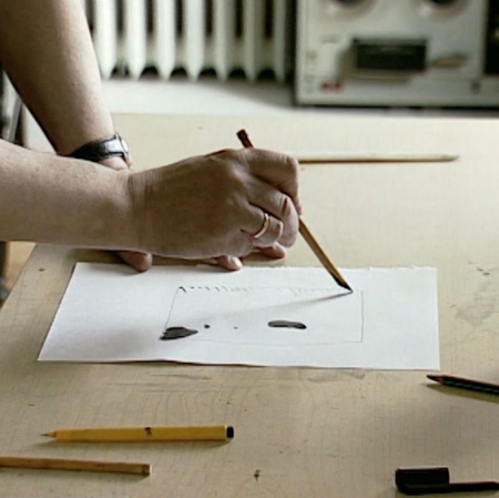  recę artysty na stole w przestrzeni pracowni, artysta trzyma w jednej rece pędzel,  rysujac na papierze. Kadr z filmu "Obraz jest moją twarzą" (1998) reż. Małgorzata Łupina, współpraca: Wojciech Leder