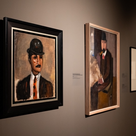 Widok wystawy. Na ścianie znajduje się szereg powieszonych obrazów. Na pierwszym planie portret mężczyzny.