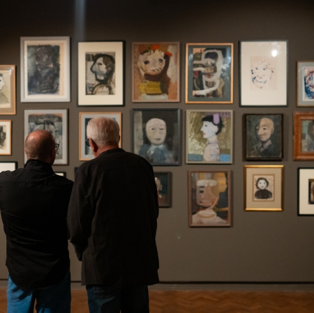 Na pierwszym planie widzimy dwie osoby odwrócone tyłem i patrzące na ścianę wypełnioną kilkunastoma pracami.