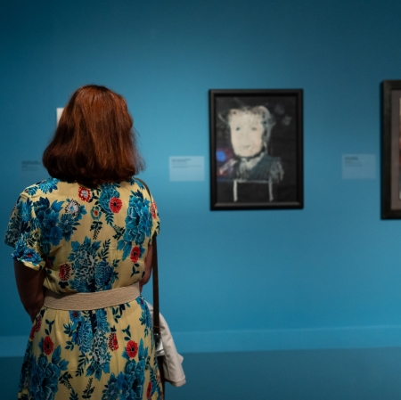 Widok wystawy. Kobieta w kwiecistej sukience stojąca w naprzeciwko kilkuportretów powieszonyh na ścianie