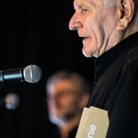 Inicjator nagrody Józef Robakowski, przemawia do mikrofonu, pod pachą trzyma kremową broszurę z białym napisem "Podanie ręki". Ma krótkie siwe włosy, w tle nieostra postać fot. HaWa