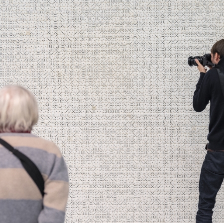 W tle ściana wypełniona kośćmi ściana. Po lewej starsza kobieta w pasiastym swetrze. Po prawo ubrany na czarno mężczyzna robiący zdjęcie.