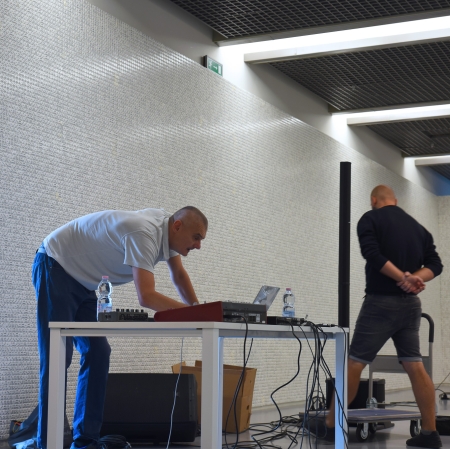 Osoba pochylona nad konsolą ustawioną na stole. W tle ściany pokryte kośćmi. Po prawo osoba stojąca z założonymi za plecami rękoma.