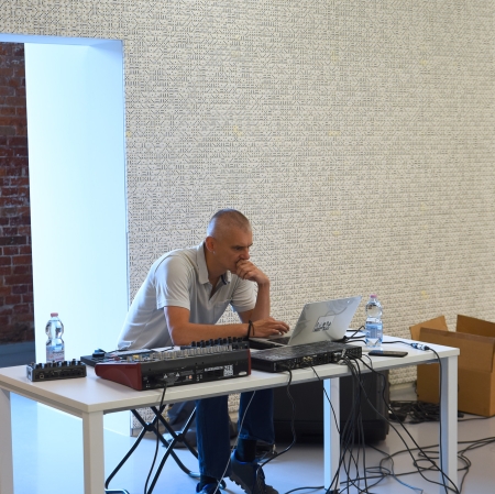 Osoba siedząca przy stole, na którym leży konsola dj-ska oraz koputer. Osoba jest pochylona nad sprzętem.