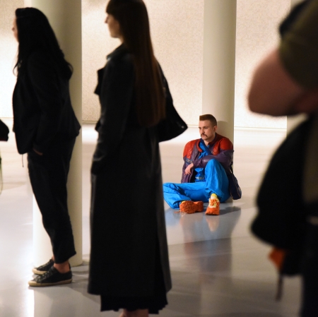 Na pierwszym planie ciemne sylwetki osób. W tle osoba siedząca na ziemi, oparta o filar. 