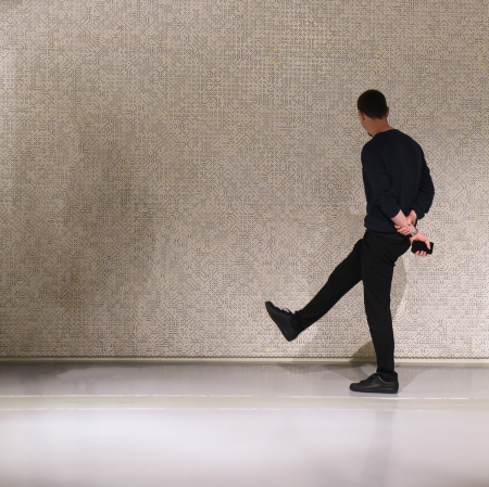 Osoba w przestrzeni wystawy unosząca nogę do góry, trzymająca splecione za plecami ręce. W tle ściany wypełnione kośćmi.