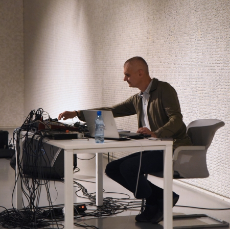 Osoba siedząca przy stole, na którym ustawiony jest komputer oraz konsola. W tle ściany wypełnione kośćmi.