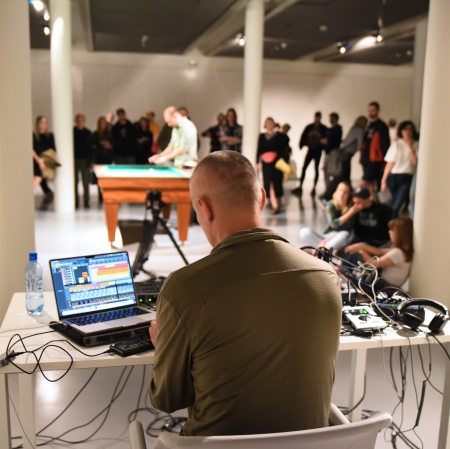 Osoba widziana z tyłu. Osoba siedzi przy stole, na którym znajduje się komputer, oraz konsola. W tle duża grupa osób słuchająca koncertu. 