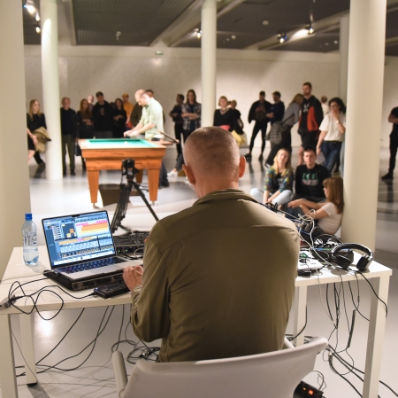 Osoba widziana z tyłu. Osoba siedzi przy stole, na którym znajduje się komputer, oraz konsola. W tle duża grupa osób słuchająca koncertu. 