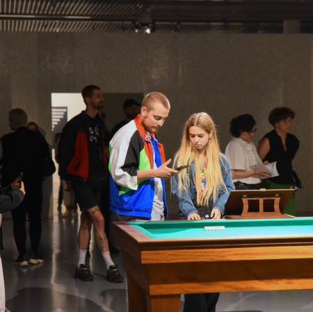 Na pierwszym planie dwie osoby rzucające kośćmi. W tle grupa osób w przestrzeni wystawy.