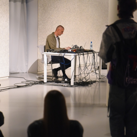 Na pierwszym planie ciemne sylwetki osób. W tle przy meżczyzna pochylony na stojącą na stole konsolą.