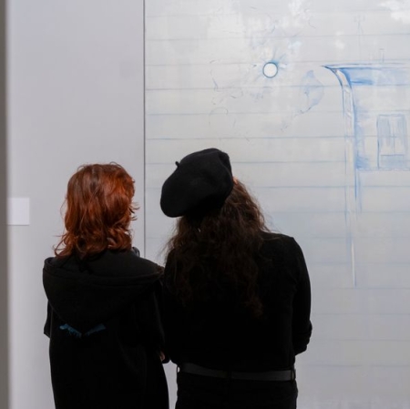 Dwie osoby odwórone tyłem, jedna z nich ma na głowie beret. Patrzą na wiszący przed nimi obraz.