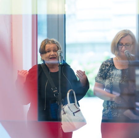 Dwie kobiety stoją przy mikrofonach. Ta stojąca od lewej strony mówi, unosząc dłonie. 