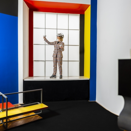osoba w goglach vr, ubrana w garnitur w kratę, stoi z uniesionymi dłońmi na parapecie. Osoba znajduje się w pomalowanej w kolory podstawowe Sali Neoplastycznej
