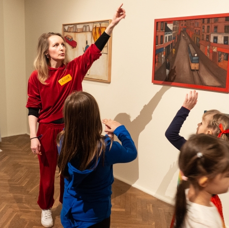 Przestrzeń wystawy. Dzieci skierowane są w stronę edukatorki, która wskazuje na obraz na ścianie.