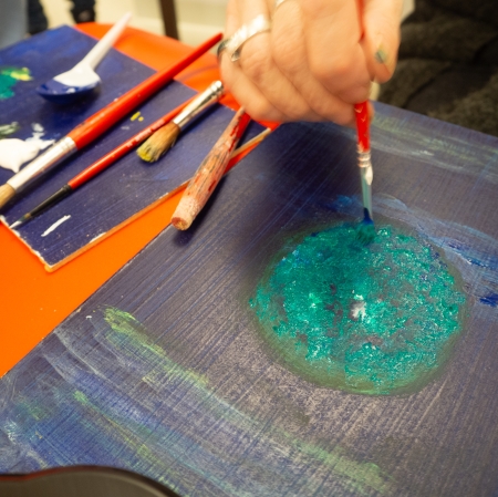 dłoń trzymająca czerwony pędzel i  malująca koło na czarnym kartonie. Na drugim planie pędzle o różnych szerokościach.