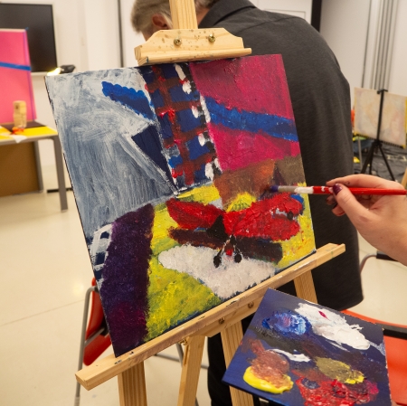 Na głównych planie abstrakcyjny obraz ustawiony na sztaludze. Po prawej dłoń trzymająca pędzel i malująca.