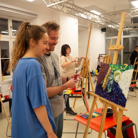 Kobieta i mężczyzna stojący na pierwszym planie przy sztaludze i rozmawiający o obrazie, który się tam znajduje.