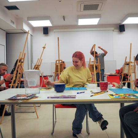 Grupa osób w różnym wieku w przestrzeni sali edukacyjnej. Jedni z nich malują siedząc przy stołach, inni przy sztalugach.